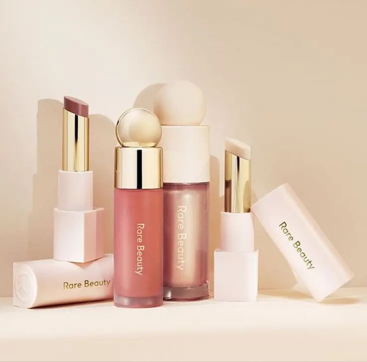 Rare beauty makeup products arranged on a soft pink background.